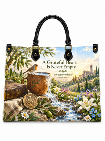 A tote bag with black handles and gold-tone hardware, featuring a detailed scenic print of a garden stream, flowers, books, a bronze mug, and a small bird, with the text 'A Grateful Heart Is Never Empty' and 'My cup overflows. Psalm 34:5.