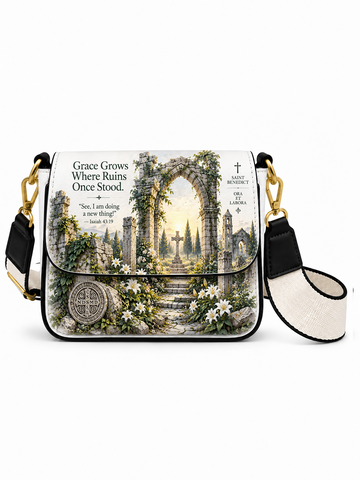 A crossbody bag with a black and white canvas strap, gold-tone hardware, and a front flap featuring a detailed religious illustration of an ancient stone archway with white lilies and the text 'Grace Grows Where Ruins Once Stood. Isaiah 45:14'.