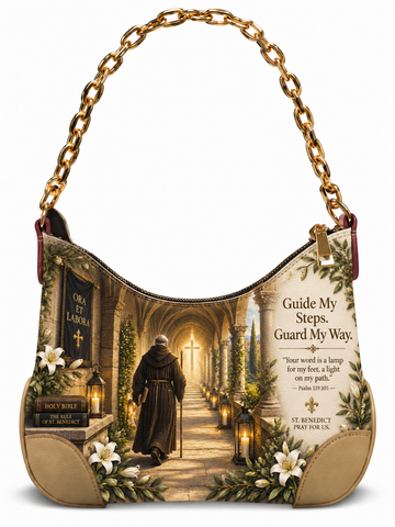 A small shoulder bag with a gold-tone chain handle, featuring a printed religious scene of a monk walking down a lit stone aisle toward a cross, with white lilies and the text 'Guide My Steps. Guard My Way.