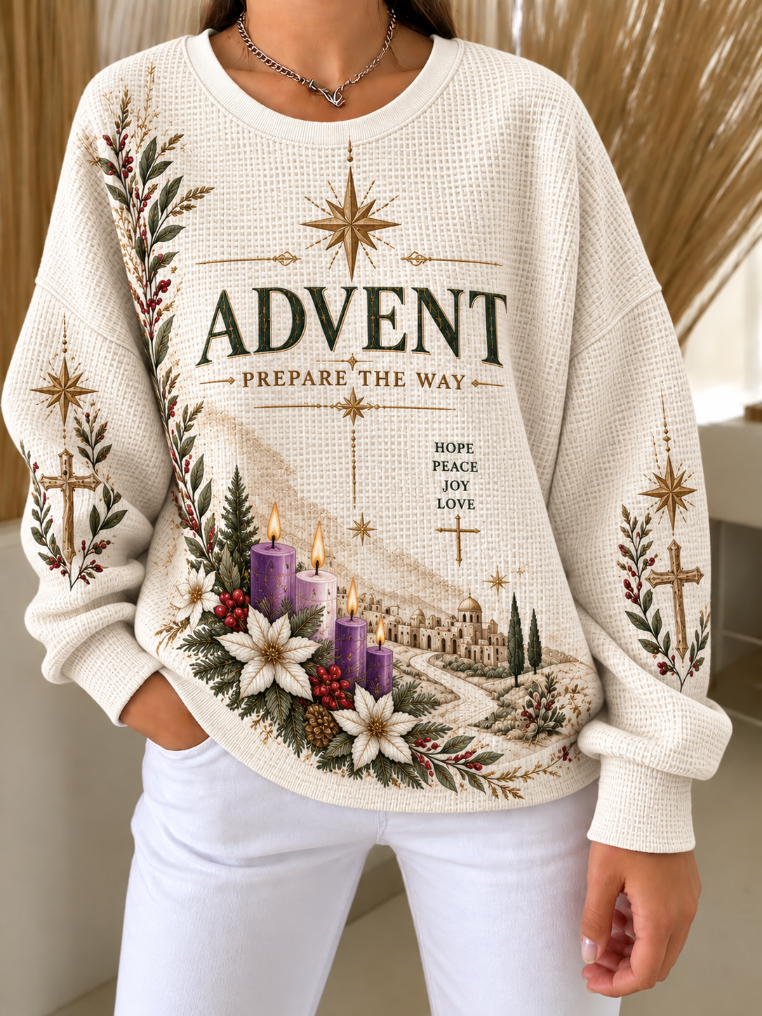 A woman wearing a cream waffle-knit sweater with a detailed Christmas Advent graphic featuring the word 'ADVENT', candles, flowers, and a small village scene, paired with white pants.