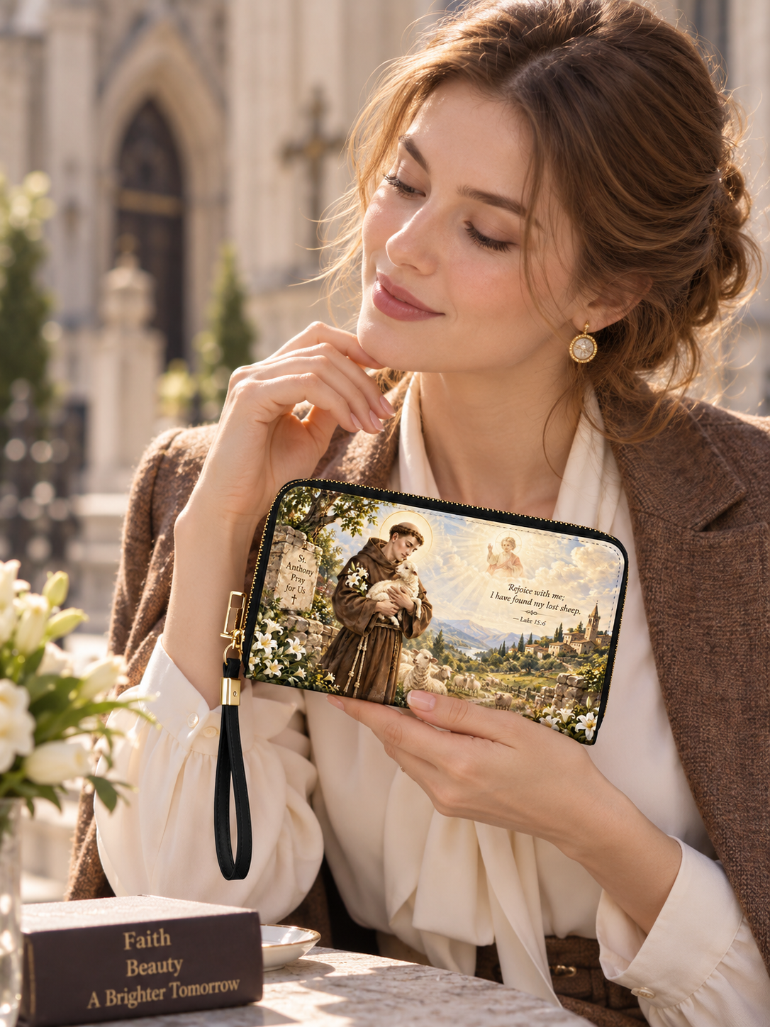 A zip-around wallet with a black wrist strap, featuring a printed religious scene of Saint Francis of Assisi holding a sheep, with white lilies, a stone sign, a shepherd, mountains, and a heavenly figure in the background. The wallet is shown on a plain white background.