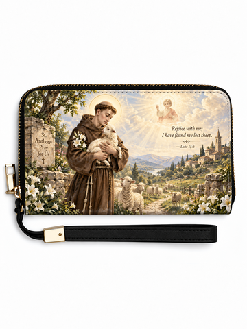 A zip-around wallet with a black wrist strap, featuring a printed religious scene of Saint Francis of Assisi holding a sheep, with white lilies, a stone sign, a shepherd, mountains, and a heavenly figure in the background. The wallet is shown on a plain white background.