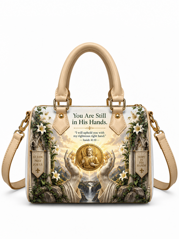 A structured handbag with beige handles and shoulder strap, featuring a detailed religious graphic print with angelic hands, a gold medallion, white flowers, and scripture text on a white background.