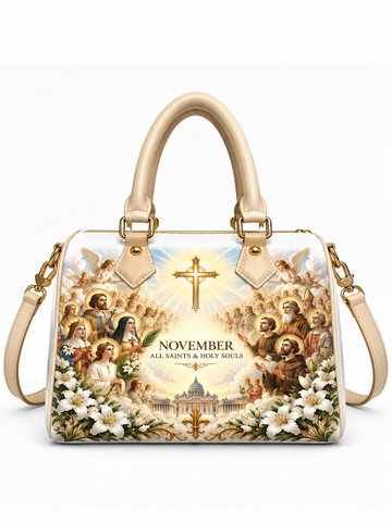 A structured cream-colored handbag with a large religious illustration printed on the front, featuring saints, angels, a radiant cross, white lilies, and the text 'NOVEMBER ALL SAINTS & HOLY SOULS.' The bag has double top handles, gold-tone hardware, and a detachable shoulder strap, shown on a clean white background.