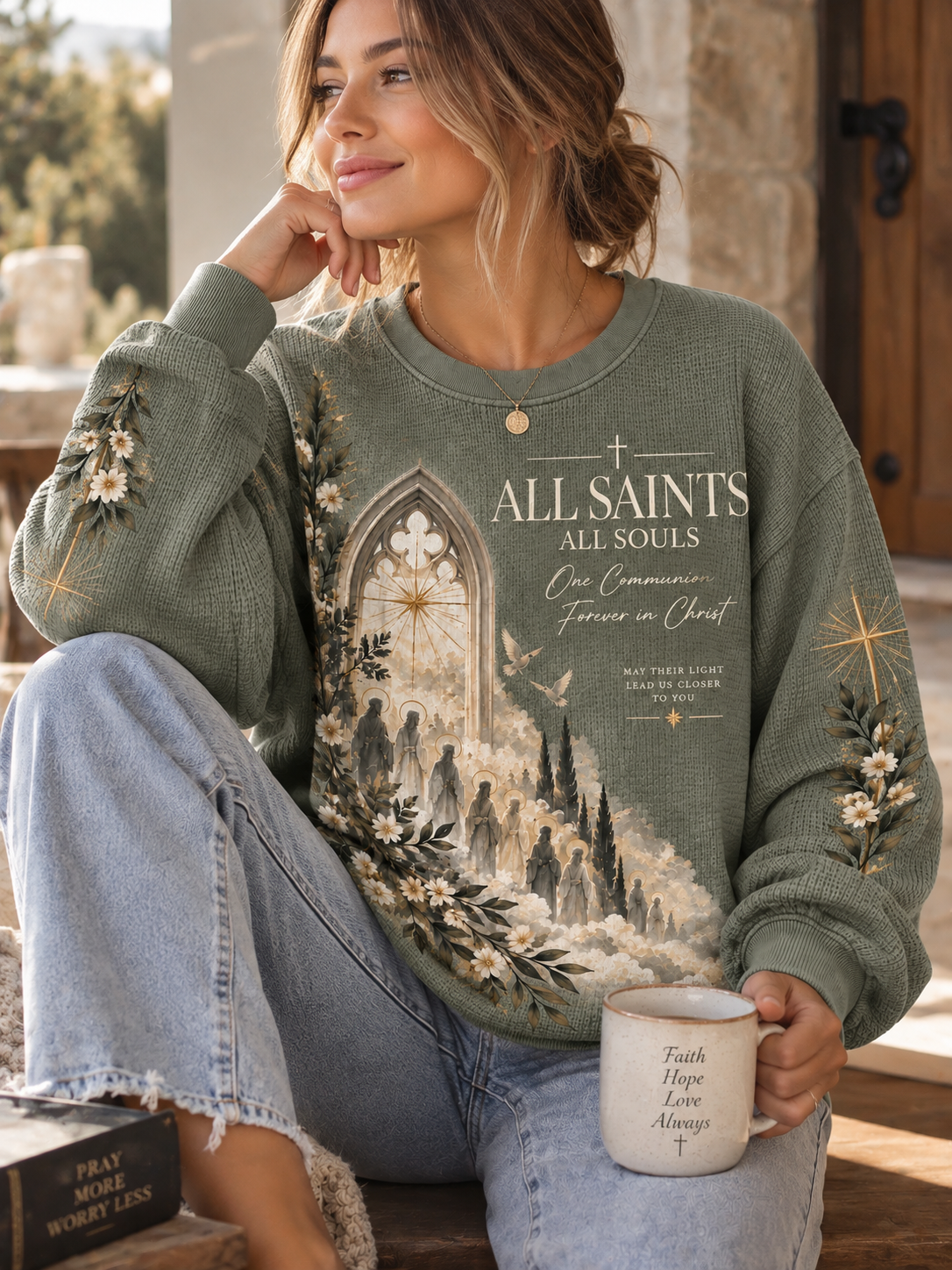 A woman wearing a sage green waffle-knit sweatshirt with a detailed graphic of a gothic cathedral and the text 'ALL SAINTS ALL SOULS' sitting on a porch and holding a white mug.
