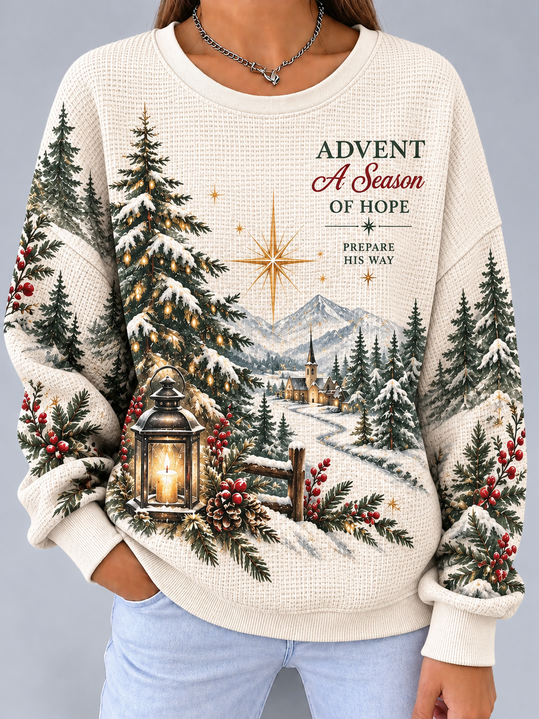 A woman wearing a cream-colored waffle-knit sweater with a detailed Christmas village scene printed on the front, featuring pine trees, mountains, a lantern, and the text 'ADVENT A Season OF HOPE'.