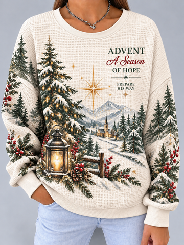 A woman wearing a cream-colored waffle-knit sweater with a detailed Christmas village scene printed on the front, featuring pine trees, mountains, a lantern, and the text 'ADVENT A Season OF HOPE'.