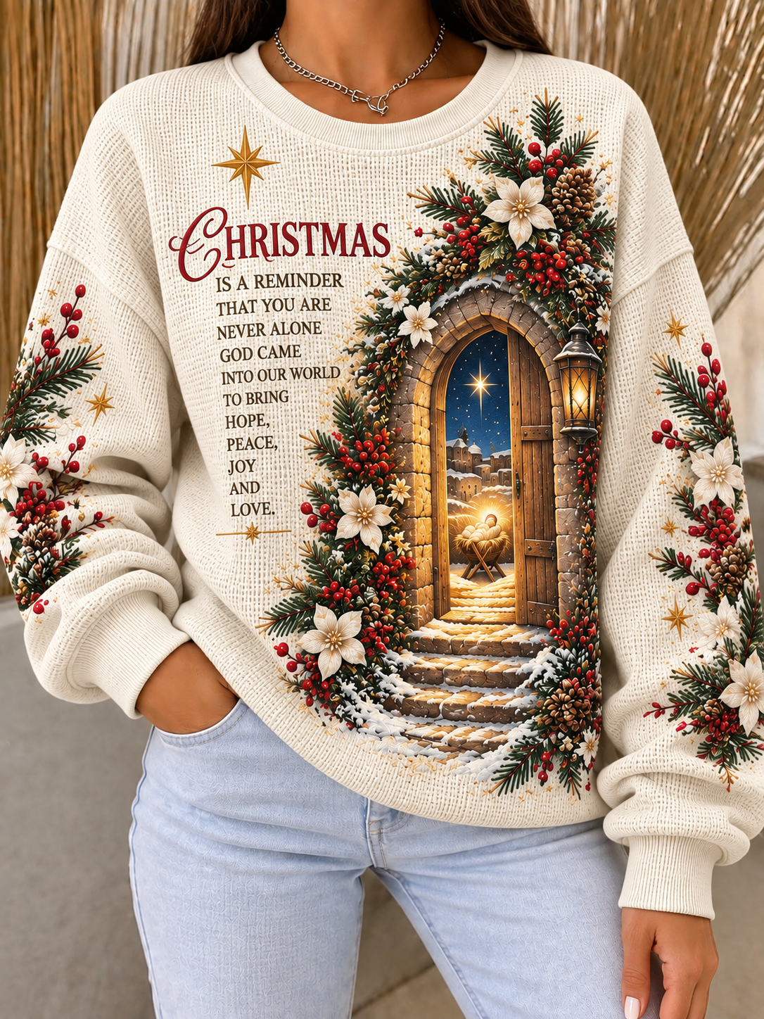 A woman wearing a cream-colored Christmas crewneck sweatshirt with a detailed graphic of an arched doorway decorated with pine garlands, red berries, and snow, alongside the text 'CHRISTMAS IS A REMINDER THAT YOU ARE NEVER ALONE.